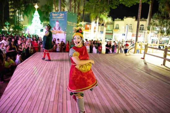 Personagem artística fantasiada em traje vermelho natalino se apresenta no palco montado no Centro de Jundiaí durante a abertura do Natal Luz 2025 da ACE, diante de um público reunido e uma árvore de Natal iluminada ao fundo.