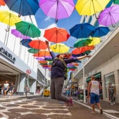 Pessoa saltando sob um corredor de guarda-chuvas coloridos na Rua Barão de Jundiaí, com pedestres caminhando ao redor e lojas ao fundo.