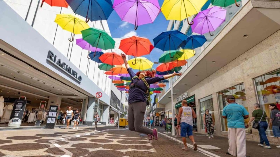 Pessoa saltando sob um corredor de guarda-chuvas coloridos na Rua Barão de Jundiaí, com pedestres caminhando ao redor e lojas ao fundo. Pessoa saltando sob um corredor de guarda-chuvas coloridos na Rua Barão de Jundiaí, com pedestres caminhando ao redor e lojas ao fundo.