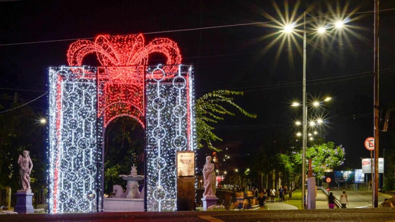 Estrutura iluminada em forma de presente gigante na entrada de espaço público de Várzea Paulista, com esculturas e fonte ao fundo.