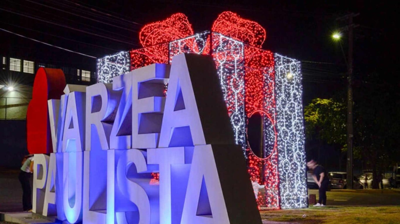 Letreiro “Várzea Paulista” iluminado ao lado de presente natalino gigante com luzes vermelhas e brancas.