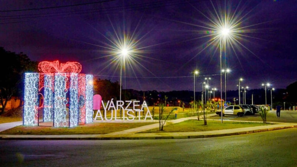 Presente natalino gigante iluminado no Parque da Família ao lado do letreiro “Várzea Paulista”, durante a noite.