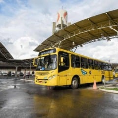 Ônibus amarelo do transporte público de Jundiaí no Terminal Vila Arens, com cobertura metálica, piso molhado e passageiros ao fundo.