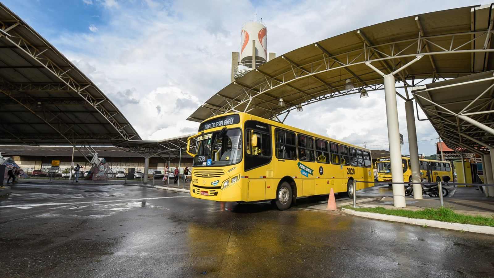 Ônibus amarelo do transporte público de Jundiaí no Terminal Vila Arens, com cobertura metálica, piso molhado e passageiros ao fundo.