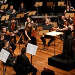Maestra rege orquestra sinf&ocirc;nica durante concerto, com m&uacute;sicos vestidos de preto tocando instrumentos de cordas e sopros em palco iluminado.