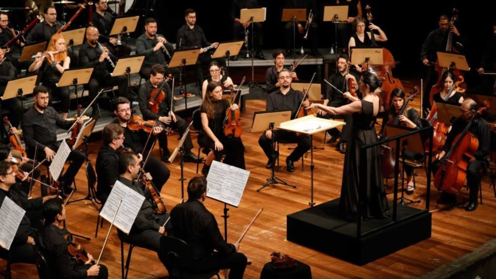 Maestra rege orquestra sinfônica durante concerto, com músicos vestidos de preto tocando instrumentos de cordas e sopros em palco iluminado. Maestra rege orquestra sinfônica durante concerto, com músicos vestidos de preto tocando instrumentos de cordas e sopros em palco iluminado.