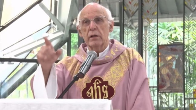 Padre Júlio Lancellotti celebra missa e fala ao microfone, vestindo paramentos litúrgicos, em igreja com vitrais ao fundo.