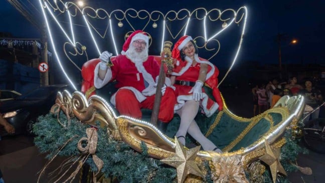 Papai Noel e Mamãe Noel em carro alegórico iluminado durante Parada Natalina em bairro de Jundiaí, com público acompanhando o desfile.