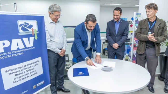 Prefeito Gustavo Martinelli assinando documento de parceria para inauguração do PAV da Receita Federal em Jundiaí, acompanhado por três autoridades, ao lado de um banner do serviço no espaço Jundiaí Empreendedora.