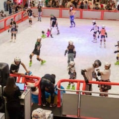 Crian&ccedil;as e adultos patinam em pista de gelo indoor cercada por grades vermelhas, dentro de shopping center, com p&uacute;blico observando ao redor.