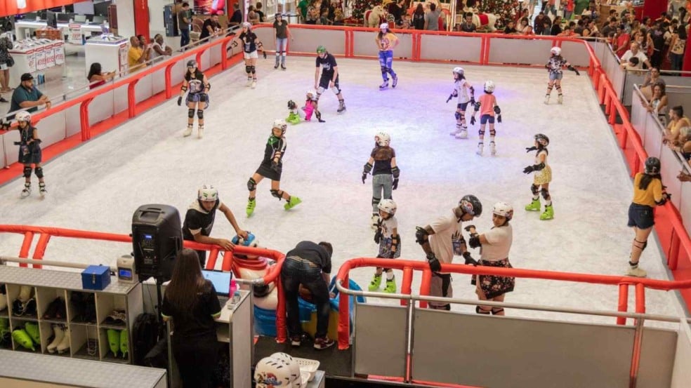Crianças e adultos patinam em pista de gelo indoor cercada por grades vermelhas, dentro de shopping center, com público observando ao redor. Crianças e adultos patinam em pista de gelo indoor cercada por grades vermelhas, dentro de shopping center, com público observando ao redor.
