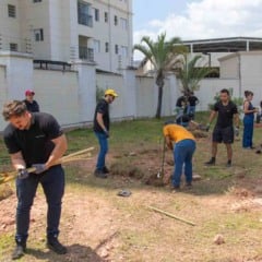 Grupo de funcionários da DHL e técnicos da Prefeitura de Jundiaí plantando mudas em área verde do bairro Eloy Chaves; pessoas utilizam pás e ferramentas enquanto preparam o solo sob céu ensolarado.