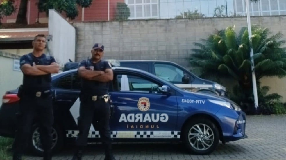 Dois guardas municipais uniformizados posam com os braços cruzados ao lado de uma viatura da Guarda Municipal de Jundiaí em área externa.