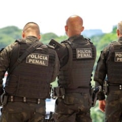 Três policiais penais uniformizados e de costas, usando coletes táticos com a inscrição “Polícia Penal”, posicionados ao ar livre com uma viatura e área verde ao fundo.