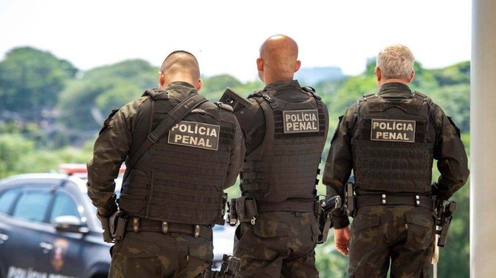 Três policiais penais uniformizados e de costas, usando coletes táticos com a inscrição “Polícia Penal”, posicionados ao ar livre com uma viatura e área verde ao fundo. Três policiais penais uniformizados e de costas, usando coletes táticos com a inscrição “Polícia Penal”, posicionados ao ar livre com uma viatura e área verde ao fundo.