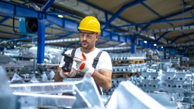 Trabalhador usando capacete e luvas de segurança inspeciona peça metálica em linha de produção dentro de uma fábrica industrial.