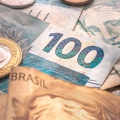 Notas e moedas do real brasileiro, com destaque para uma c&eacute;dula de R$ 100, simbolizando pagamento de benef&iacute;cios e valores financeiros.