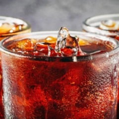 Copo de refrigerante escuro com gelo e condensação na superfície, em close, ao lado de outras bebidas semelhantes.