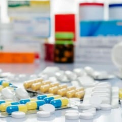 Comprimidos e c&aacute;psulas de diferentes cores e formatos espalhados sobre uma mesa, com frascos e caixas de medicamentos desfocados ao fundo.&rdquo;