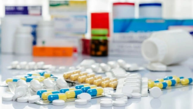 Comprimidos e cápsulas de diferentes cores e formatos espalhados sobre uma mesa, com frascos e caixas de medicamentos desfocados ao fundo.”
