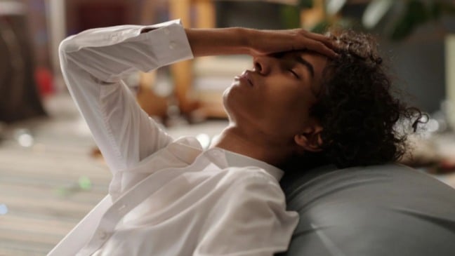 Pessoa deitada com a mão na testa e expressão de mal-estar, em ambiente desorganizado, representando sintomas de ressaca após exagero na bebida.