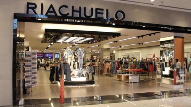 Fachada de loja da Riachuelo em shopping, com manequins, araras de roupas e ambiente interno amplo e bem iluminado.