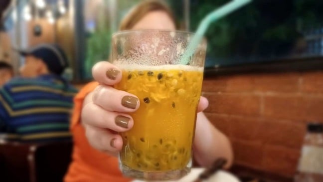 Pessoa segurando um copo de bebida alcoólica amarela com gelo e canudo, em ambiente de confraternização.