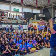 Palestrante falando ao microfone no gin&aacute;sio do Sesi Jundia&iacute; diante de dezenas de estudantes sentados, todos caracterizados com uniformes das equipes de rob&oacute;tica.