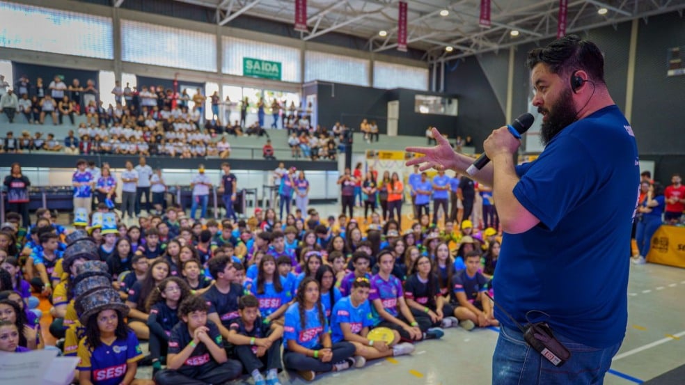 Palestrante falando ao microfone no ginásio do Sesi Jundiaí diante de dezenas de estudantes sentados, todos caracterizados com uniformes das equipes de robótica. Palestrante falando ao microfone no ginásio do Sesi Jundiaí diante de dezenas de estudantes sentados, todos caracterizados com uniformes das equipes de robótica.