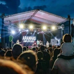 Palco iluminado da Festa da Uva à noite com a banda Aranhas Amplificadas se apresentando diante de uma grande plateia; uma criança observa o show nos ombros de um adulto.