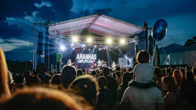 Palco iluminado da Festa da Uva à noite com a banda Aranhas Amplificadas se apresentando diante de uma grande plateia; uma criança observa o show nos ombros de um adulto.
