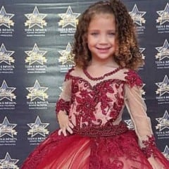 Menina de 4 anos, Pietra Hasten, usa vestido vermelho bordado e sorri diante do painel do concurso Miss São Paulo Teen Infantil.