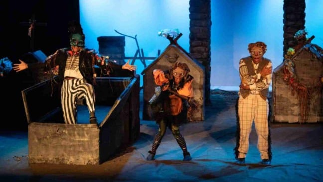 Atores da peça “R.I.P. – Uma história de amor” em cena sobre um palco ambientado como cemitério, usando máscaras e figurinos coloridos; um ator sai de um caixão cenográfico, uma atriz se agacha em pose cômica e outro ator demonstra surpresa, sob iluminação azulada.