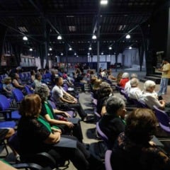 Público sentado no Espaço Expressa, em Jundiaí, assistindo à fala de um apresentador diante do palco, com vagões antigos ao fundo.
