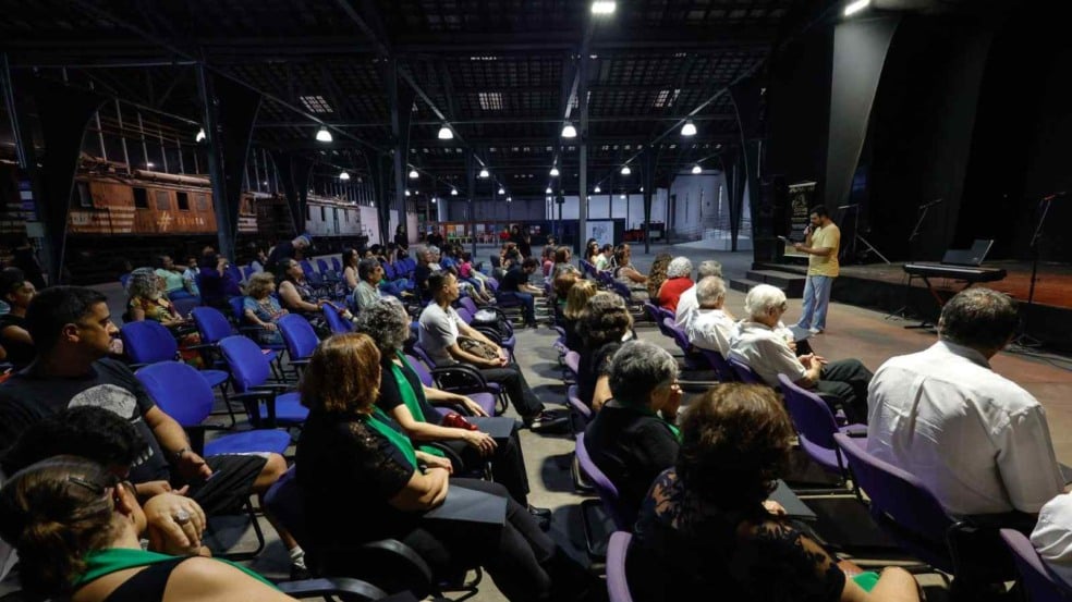 Público sentado no Espaço Expressa, em Jundiaí, assistindo à fala de um apresentador diante do palco, com vagões antigos ao fundo. Público sentado no Espaço Expressa, em Jundiaí, assistindo à fala de um apresentador diante do palco, com vagões antigos ao fundo.