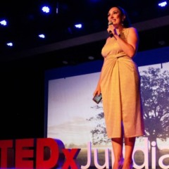 Mulher em vestido claro fala ao microfone no palco do TEDxJundia&iacute;, com o logo do evento ao lado e proje&ccedil;&atilde;o de uma &aacute;rvore ao fundo.