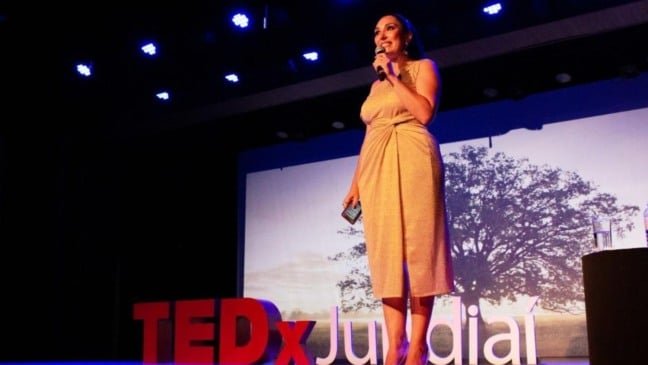 Mulher em vestido claro fala ao microfone no palco do TEDxJundiaí, com o logo do evento ao lado e projeção de uma árvore ao fundo.