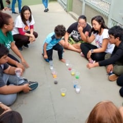 Grupo de crianças, adolescentes e adultos sentados em roda no chão, participando de uma atividade com copos cheios de bolinhas coloridas na Teia Down Jundiaí. O ambiente é externo e a imagem destaca interação, inclusão e acolhimento.