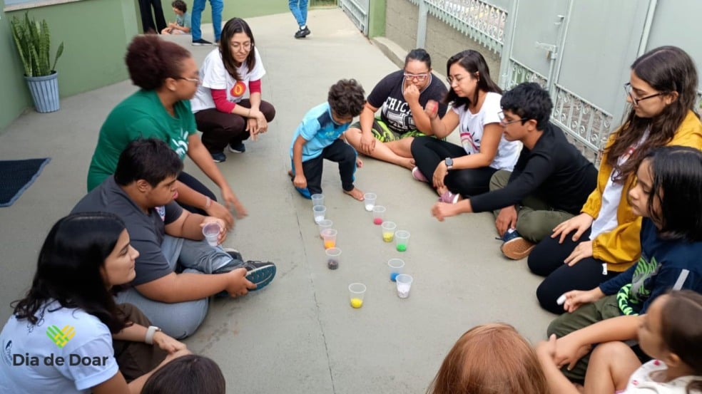 Grupo de crianças, adolescentes e adultos sentados em roda no chão, participando de uma atividade com copos cheios de bolinhas coloridas na Teia Down Jundiaí. O ambiente é externo e a imagem destaca interação, inclusão e acolhimento.