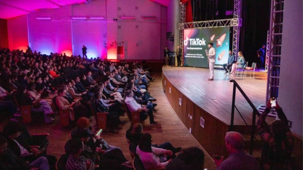 Auditório lotado durante evento do TikTok em Jundiaí, com público atento à palestra no palco iluminado, onde um apresentador fala diante de telão da plataforma. Auditório lotado durante evento do TikTok em Jundiaí, com público atento à palestra no palco iluminado, onde um apresentador fala diante de telão da plataforma.