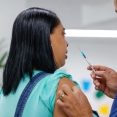 Profissional de sa&uacute;de segurando uma seringa enquanto se prepara para aplicar vacina no bra&ccedil;o de uma mulher em um ambiente de atendimento.