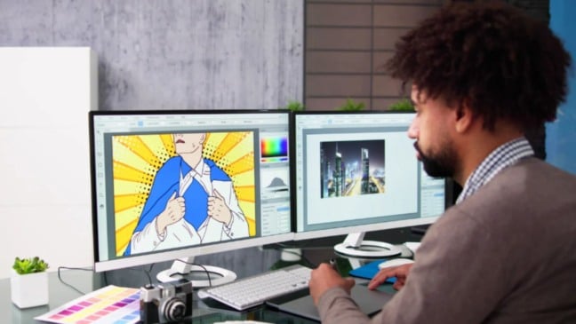 Designer trabalhando em mesa com dois monitores exibindo uma arte pop art e uma imagem de cidade, acompanhado de câmera, paleta de cores e acessórios.