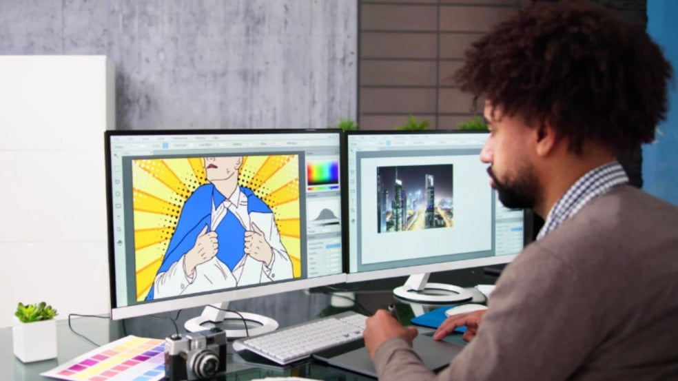 Designer trabalhando em mesa com dois monitores exibindo uma arte pop art e uma imagem de cidade, acompanhado de câmera, paleta de cores e acessórios.