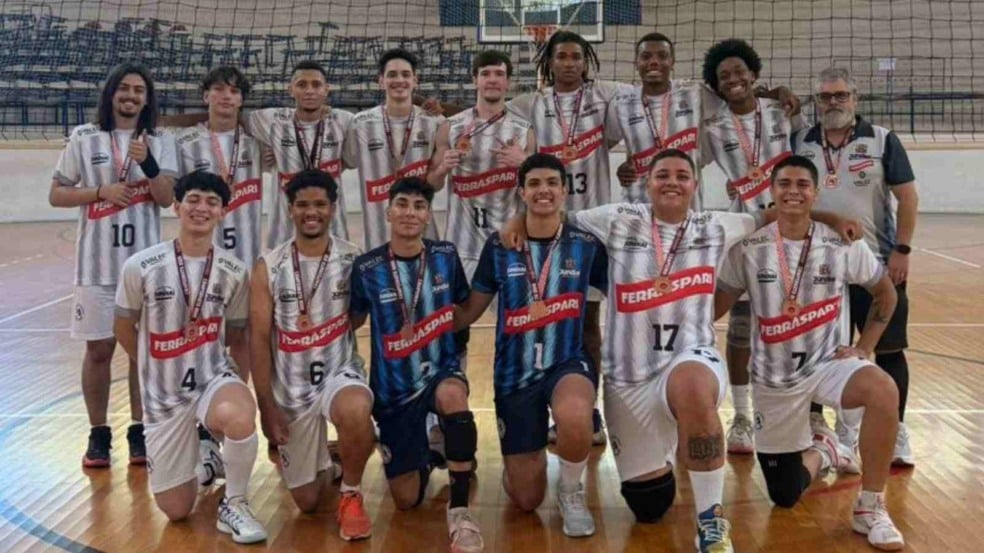 Atletas do vôlei masculino sub-21 de Jundiaí posam em quadra, usando uniformes branco e azul, mostrando medalhas de bronze ao lado do técnico. Atletas do vôlei masculino sub-21 de Jundiaí posam em quadra, usando uniformes branco e azul, mostrando medalhas de bronze ao lado do técnico.
