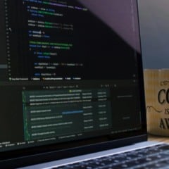 Notebook com c&oacute;digo de programa&ccedil;&atilde;o aberto na tela, ao lado de uma caneca de caf&eacute; com a frase &ldquo;This coffee is making me awesome&rdquo;, sobre uma mesa de madeira.