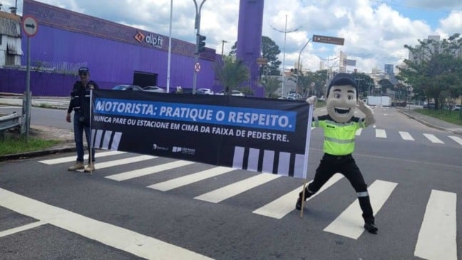 Agentes de trânsito e mascote Vaguinho seguram faixa educativa sobre respeito à faixa de pedestres durante ação em rua de Jundiaí.