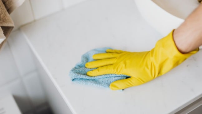 Pessoa com luvas amarelas limpando uma superfície branca com pano azul durante serviço de limpeza.