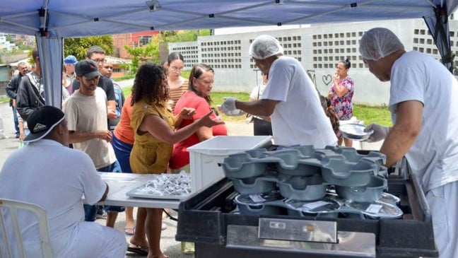 Servidores do Bom Prato Móvel distribuem marmitas a moradores em fila durante atendimento comunitário em bairro de Várzea Paulista.