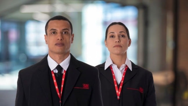 Dois vigilantes do Grupo GR, um homem e uma mulher, usando uniforme social com crachá da empresa, em ambiente corporativo.