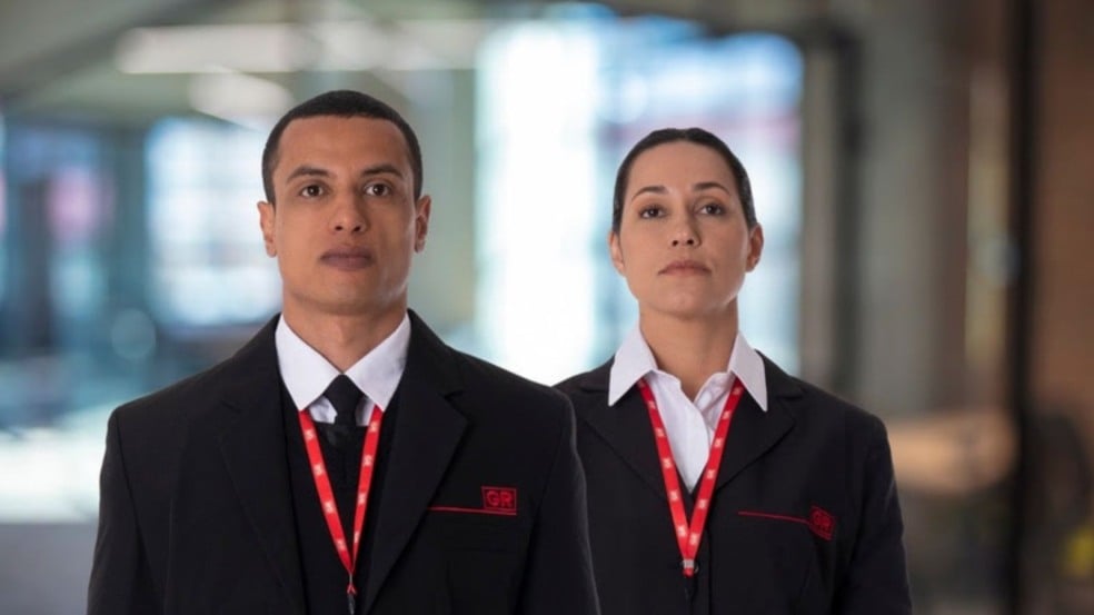 Dois vigilantes do Grupo GR, um homem e uma mulher, usando uniforme social com crachá da empresa, em ambiente corporativo. Dois vigilantes do Grupo GR, um homem e uma mulher, usando uniforme social com crachá da empresa, em ambiente corporativo.